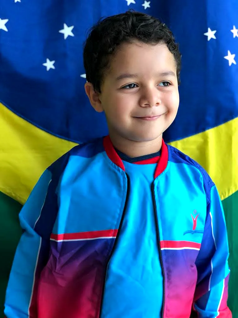 O mesmo aluno, hoje, de jaqueta do uniforme em frente à bandeira do Brasil