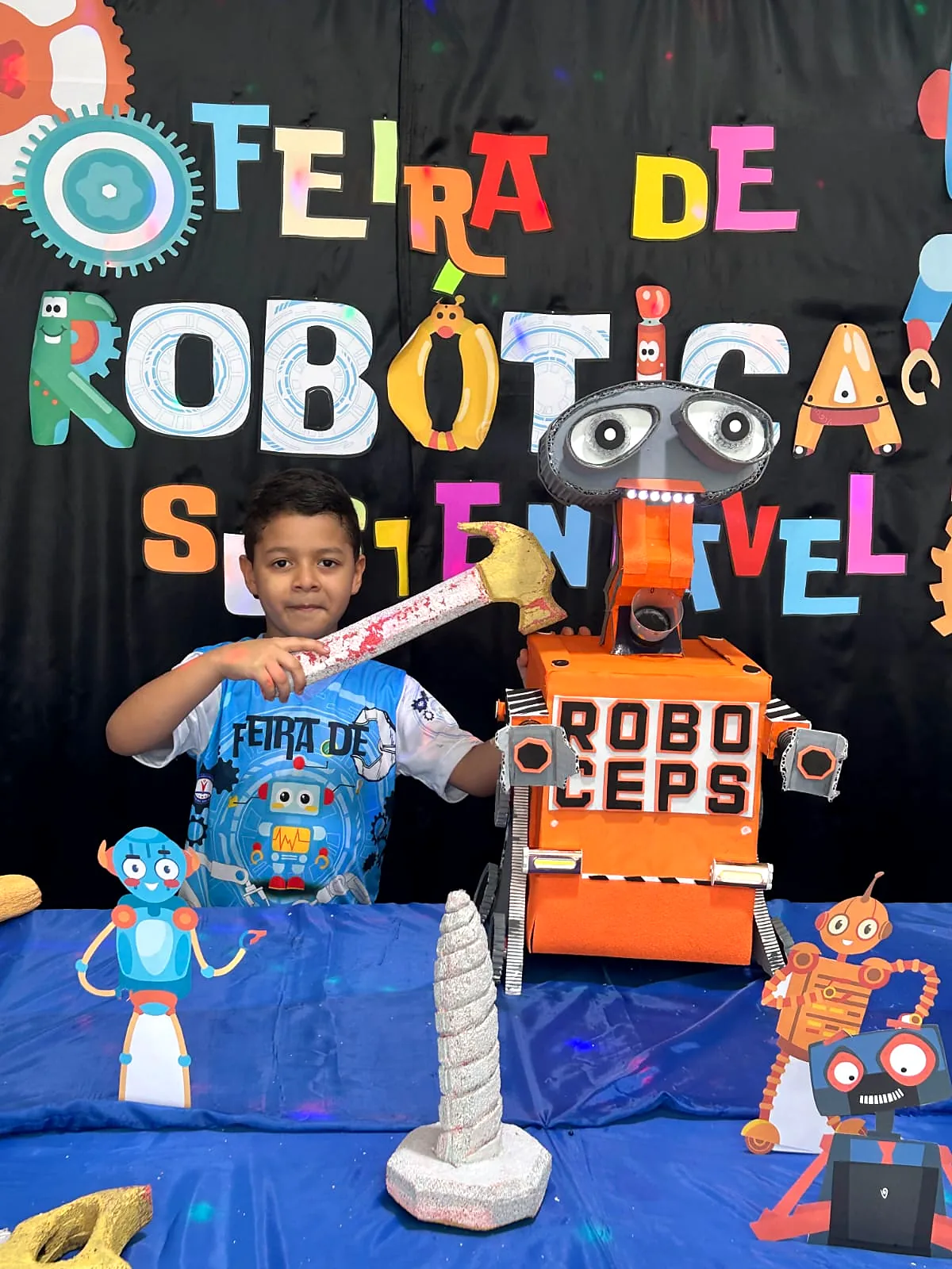 Aluno ao lado de robôs de sucata na Feira de Robótica Sustentável